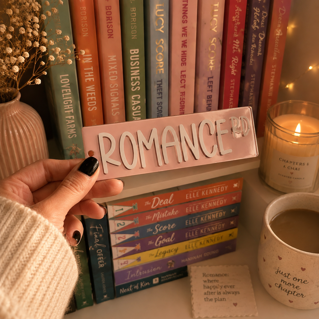 A hand with black nail polish holds a pink rectangular sign that reads 'ROMANCE 20' in white letters, positioned in front of a shelf of colorful romance novels. A lit candle, a mug, and dried flowers are visible in the warm, cozy setting.