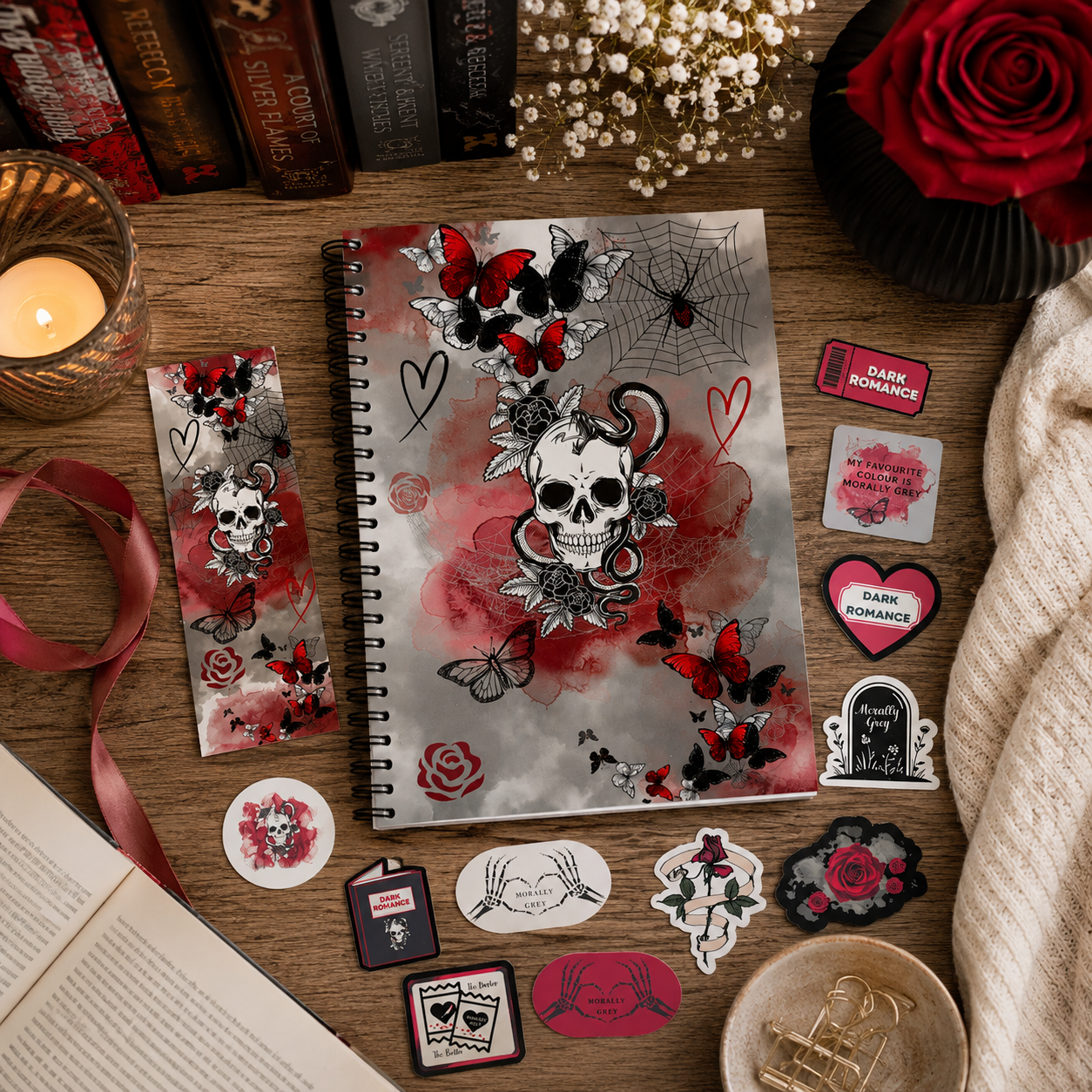 A flat-lay display of dark romance-themed stationery, featuring a spiral notebook and matching sticker/bookmark set with skull, rose, spider, and heart designs, arranged on a wooden tabletop with books, a candle, ribbon, and a cream knit sweater.