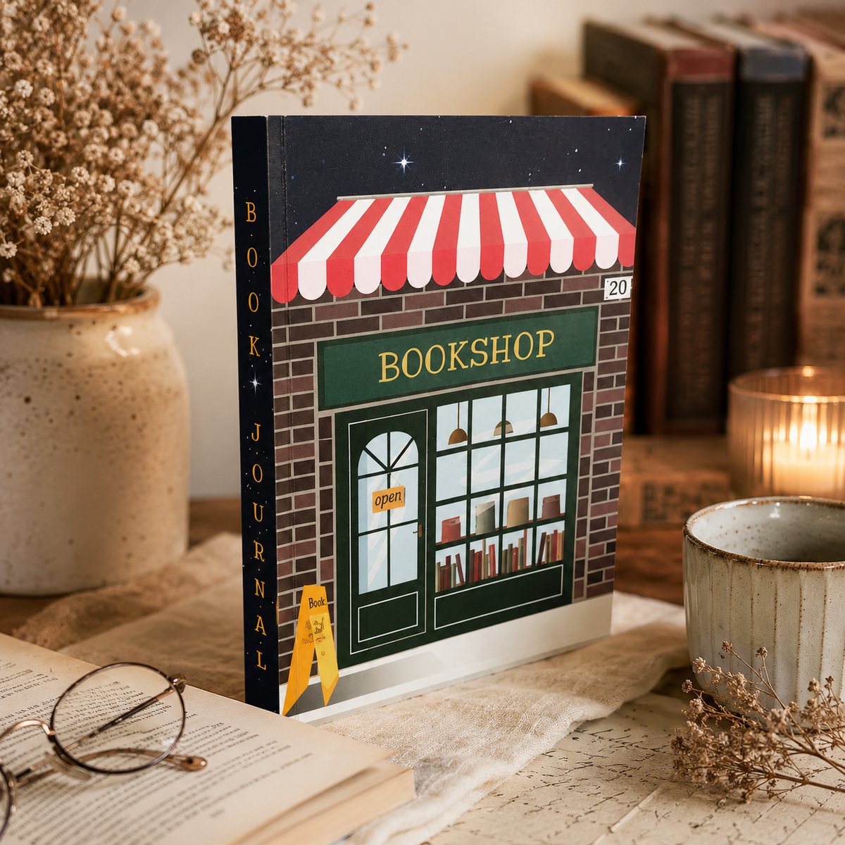 A paperback book journal titled 'Bookshop' styled on a tabletop with an open book, glasses, dried flowers, and a warm candle in the background. The cover features an illustration of a brick bookshop with a red and white striped awning.