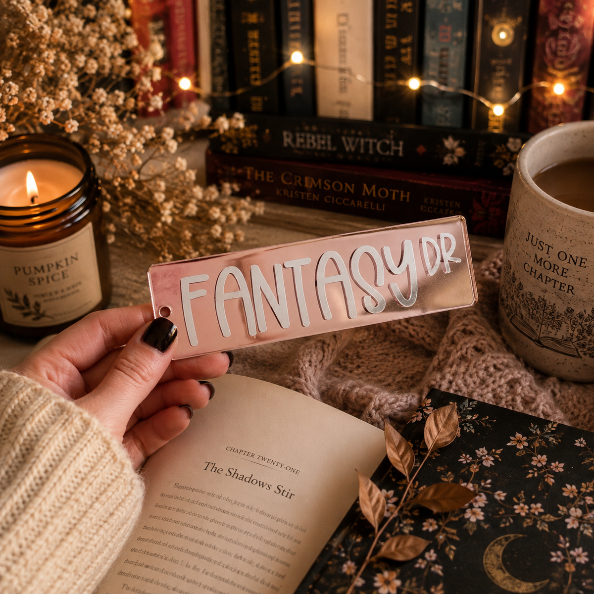 A hand with black nail polish holds a clear acrylic bookmark featuring the text 'FANTASY DR' in white. The scene is styled with cozy, fantasy-themed props including a candle, books, a mug, and an open book.