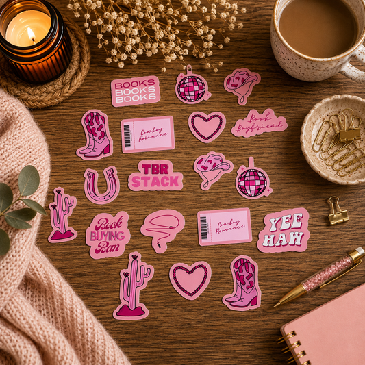 A flat-lay arrangement of pink and magenta sticker designs on a wooden table, surrounded by cozy props including a lit candle, a cup of coffee, dried flowers, a pink knit blanket, a pen, and a notebook.