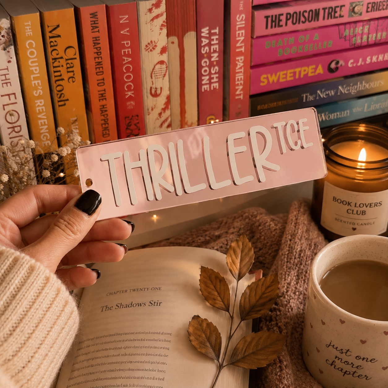 A hand with black nail polish holds a pink rectangular bookend featuring the word 'THRILLER' in raised white letters. The background is a cozy, styled bookshelf scene with books, a lit candle, a mug, and an open book.