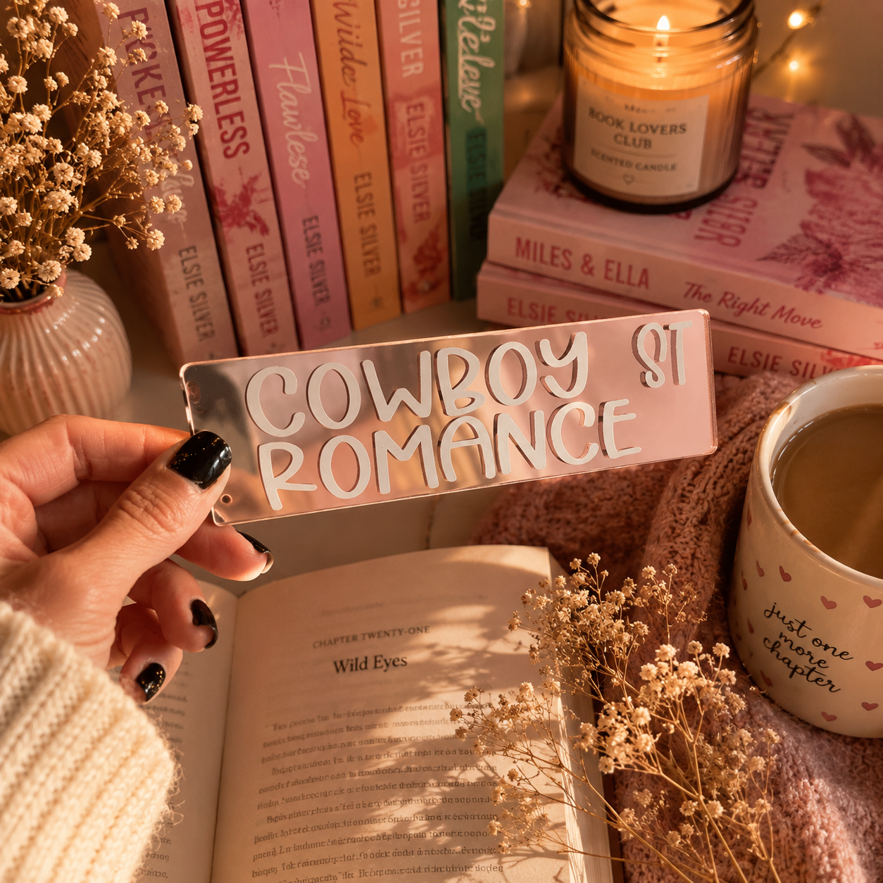 A hand with black nail polish holds a rectangular metal sign that reads 'COWBOY ST ROMANCE' in raised white letters. The sign has a pink glittery background and is photographed in a cozy, book-themed setting with books, a candle, a mug, and dried flowers.
