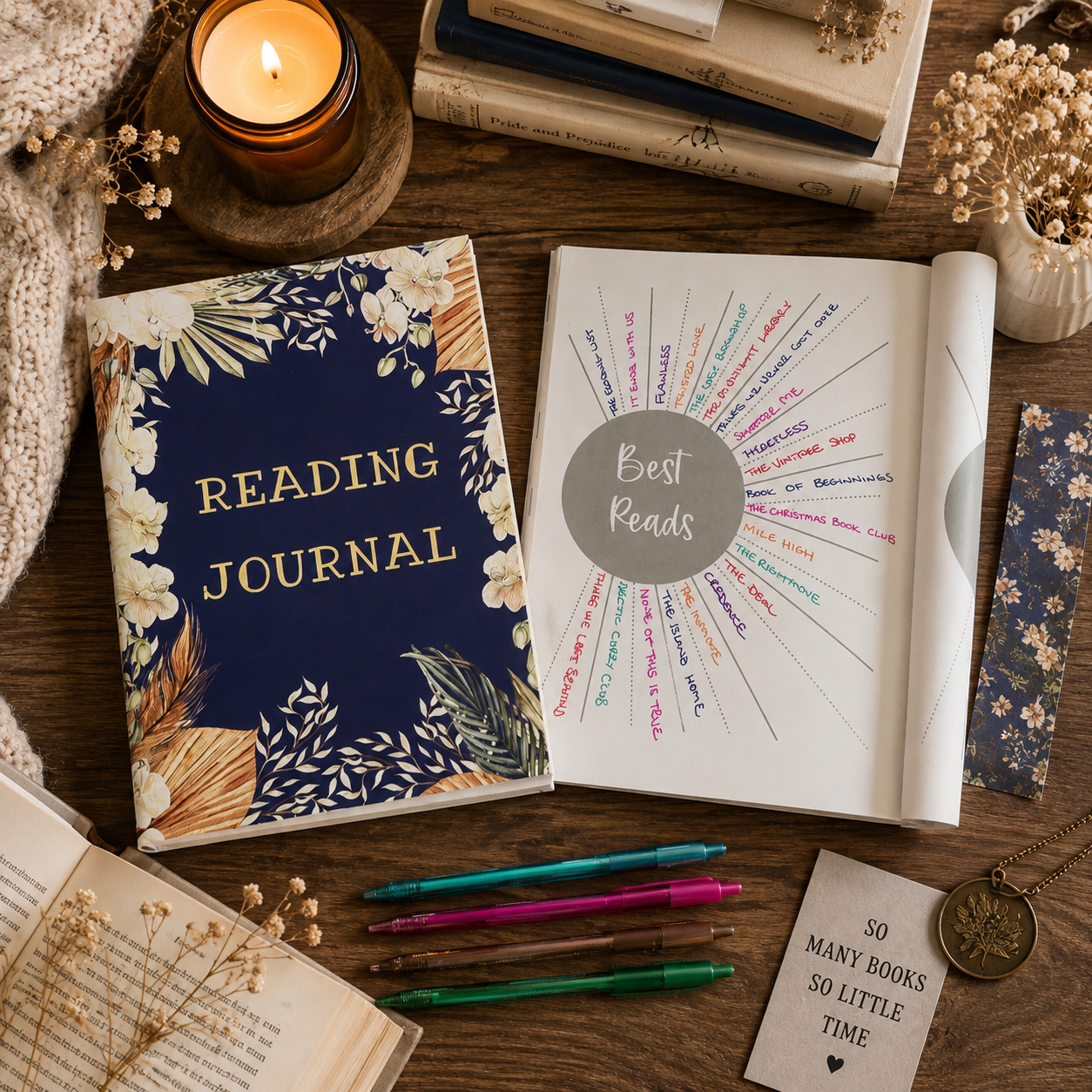 A styled flat-lay of reading materials on a wooden table, featuring a navy floral 'READING JOURNAL' with gold lettering, an open white notebook with a circular 'Best Reads' chart, colorful pens, books, a candle, dried flowers, and a small card.