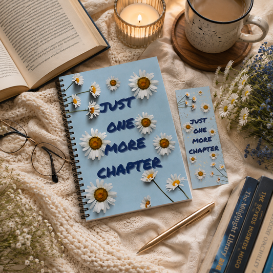 A light blue spiral notebook with daisy decorations and the words 'JUST ONE MORE CHAPTER' is styled on a cream lace blanket with an open book, glasses, a candle, a mug, and other cozy reading props. In the style of Lucy Score