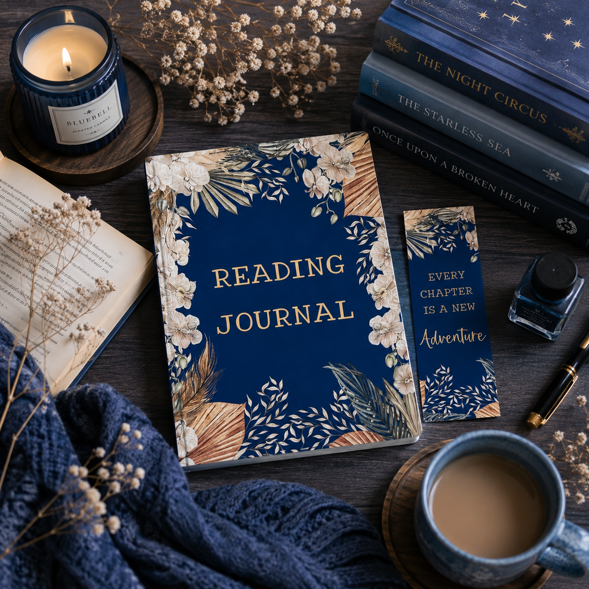 A navy blue reading journal with gold lettering and floral illustrations, styled on a dark wooden table with a matching bookmark, books, a candle, a cup of coffee, and dried flowers.