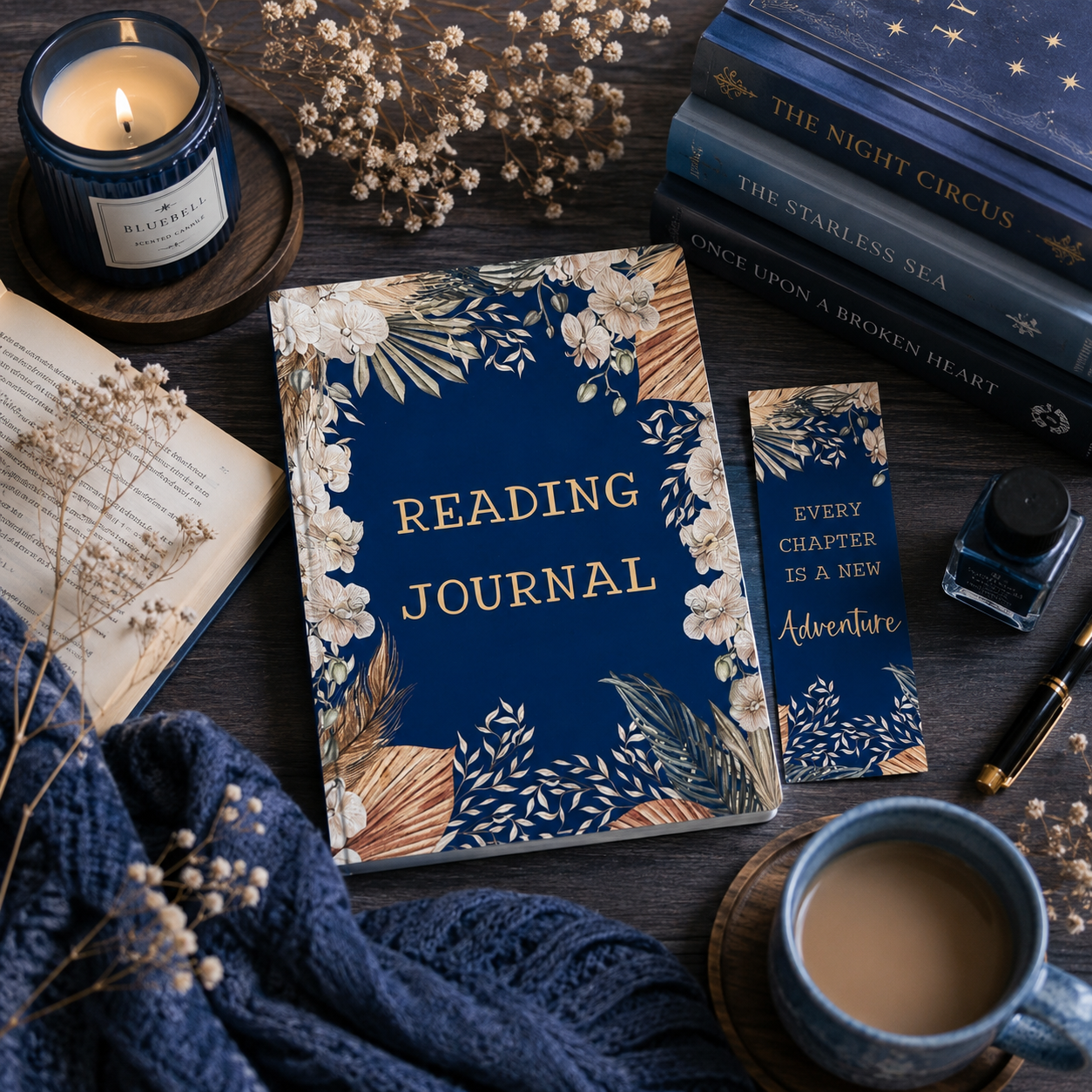 A navy blue reading journal with gold lettering and floral illustrations, styled on a dark wooden table with a matching bookmark, books, a candle, a cup of coffee, and dried flowers.