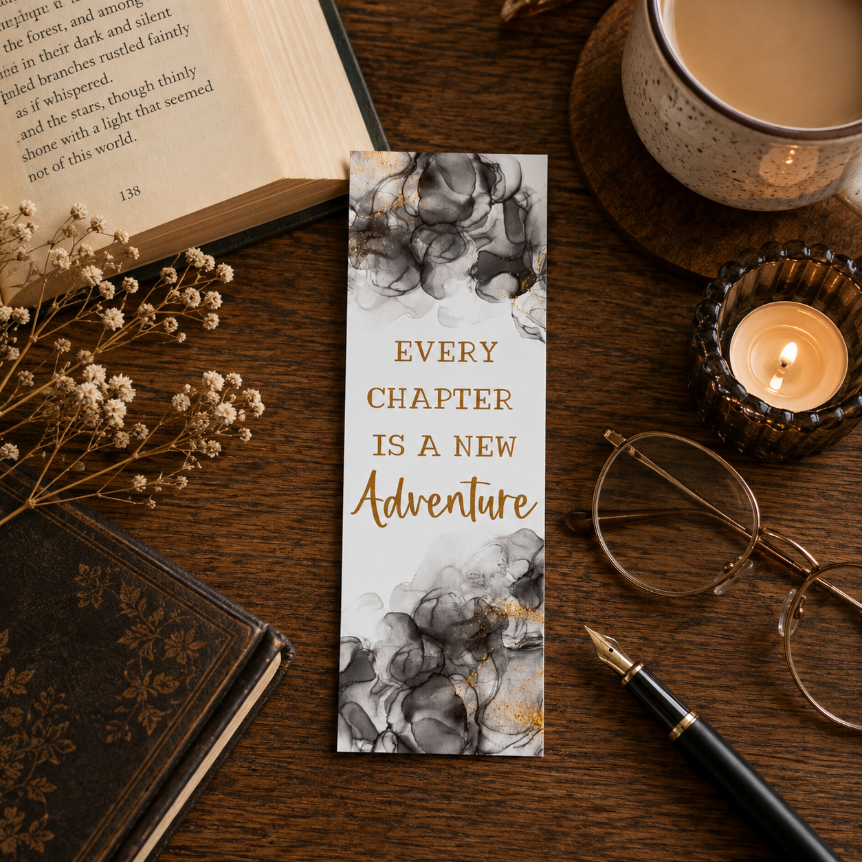 A vertical rectangular bookmark or book divider with a white background, gray and black marble-style floral clouds at the top and bottom, and the phrase 'EVERY CHAPTER IS A NEW Adventure' in gold lettering, placed on a wooden desk with books, glasses, a candle, and a pen around it.