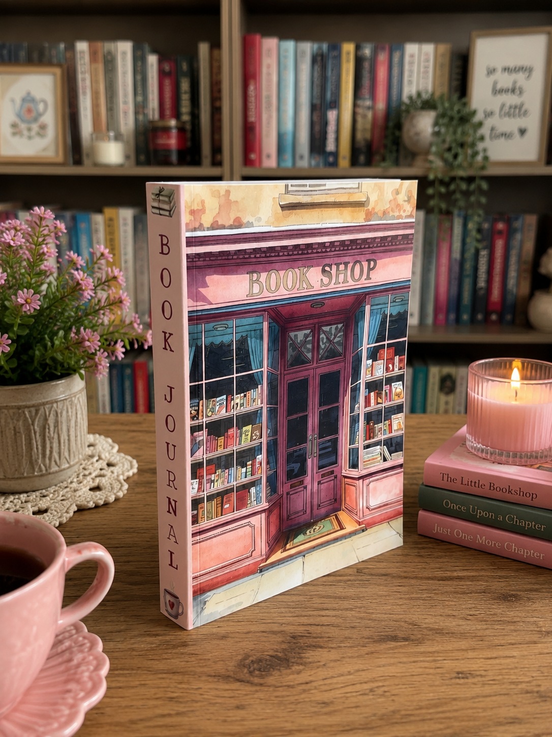 A bookshop Book Journal standing upright on a wooden table, featuring an illustration of a pink bookshop storefront on the cover. The scene is styled with a pink teacup, a pink candle, a small potted plant, and a stack of books, with a bookshelf filled with books in the background.