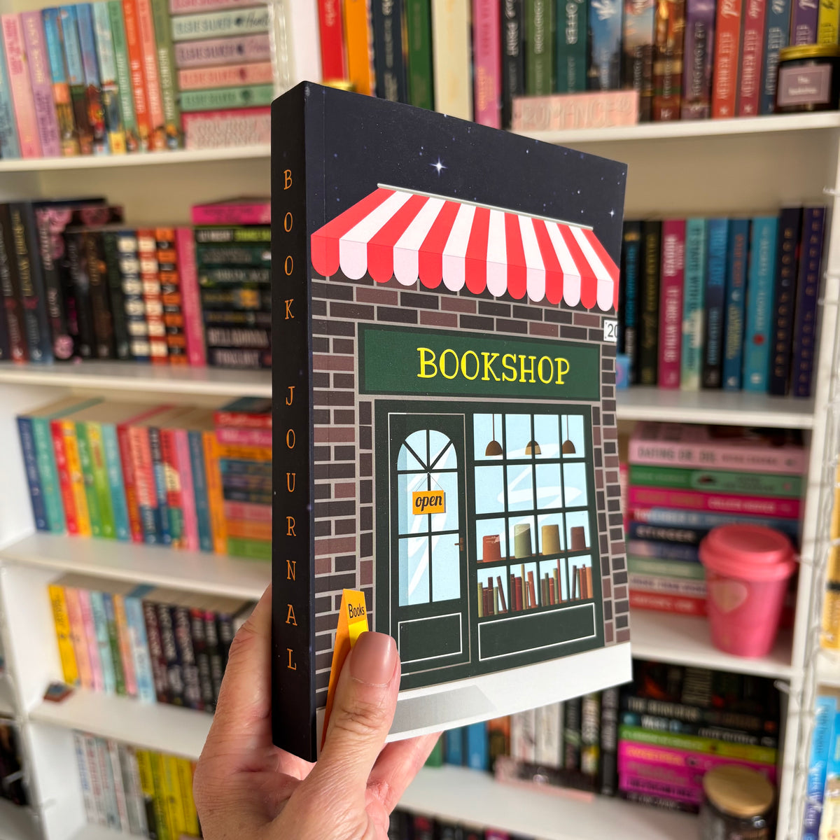 A hand holding a black bookshop-themed reading log journal with a red and white striped awning design on the cover, in front of a bookshelf filled with various books.