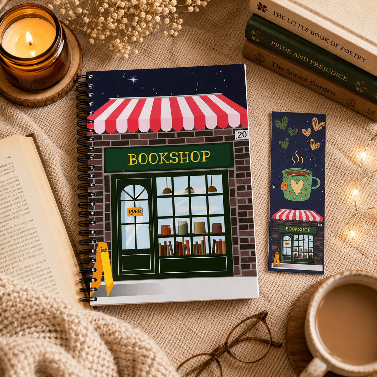 A spiral-bound reading journal and matching bookmark set featuring a cosy bookshop illustration, styled on a beige fabric surface with a candle, open book, mug of coffee, glasses, and decorative books.