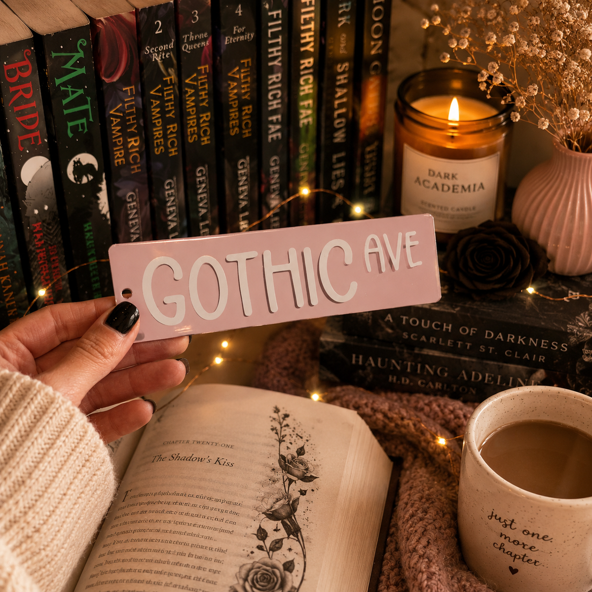 A hand with black nail polish holds a pink bookmark sign reading 'GOTHIC AVE' in front of a cozy bookshelf scene with books, a lit candle, an open book, a mug, and warm string lights.
