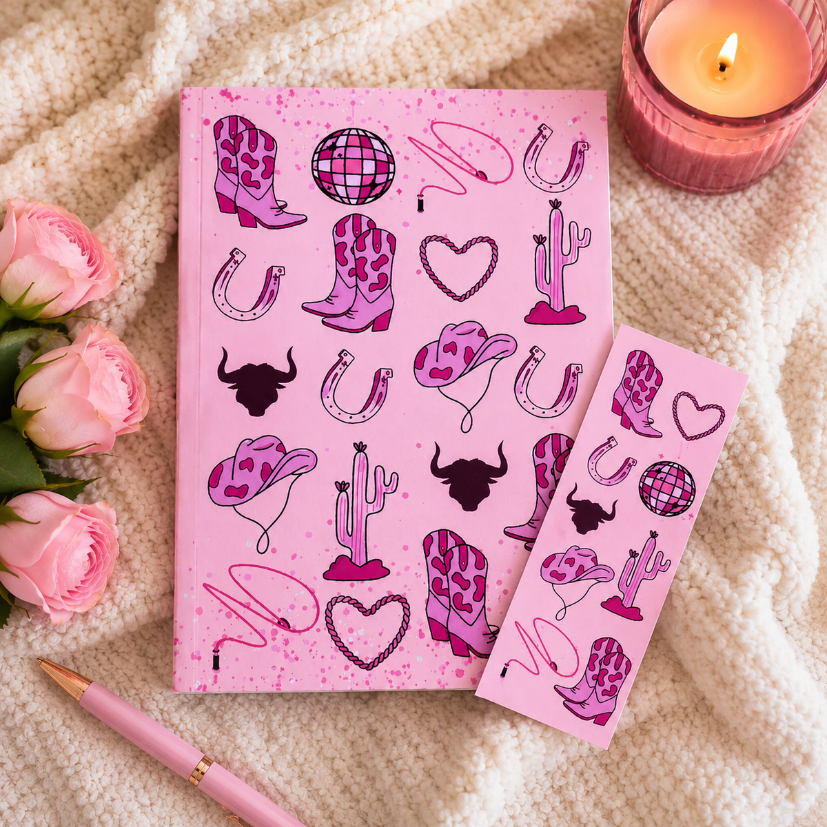 A pink book journal and matching bookmark set decorated with western-themed illustrations including cowboy boots, horseshoes, cacti, and hearts, styled on a cream knit blanket with pink roses and a lit candle.