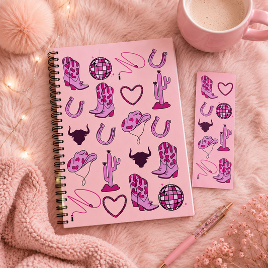 A pink spiral reading journal and matching bookmark featuring western-themed illustrations including cowboy boots, horseshoes, cacti, hearts, and disco balls, styled on a fluffy cream surface with a mug, pen, and string lights.