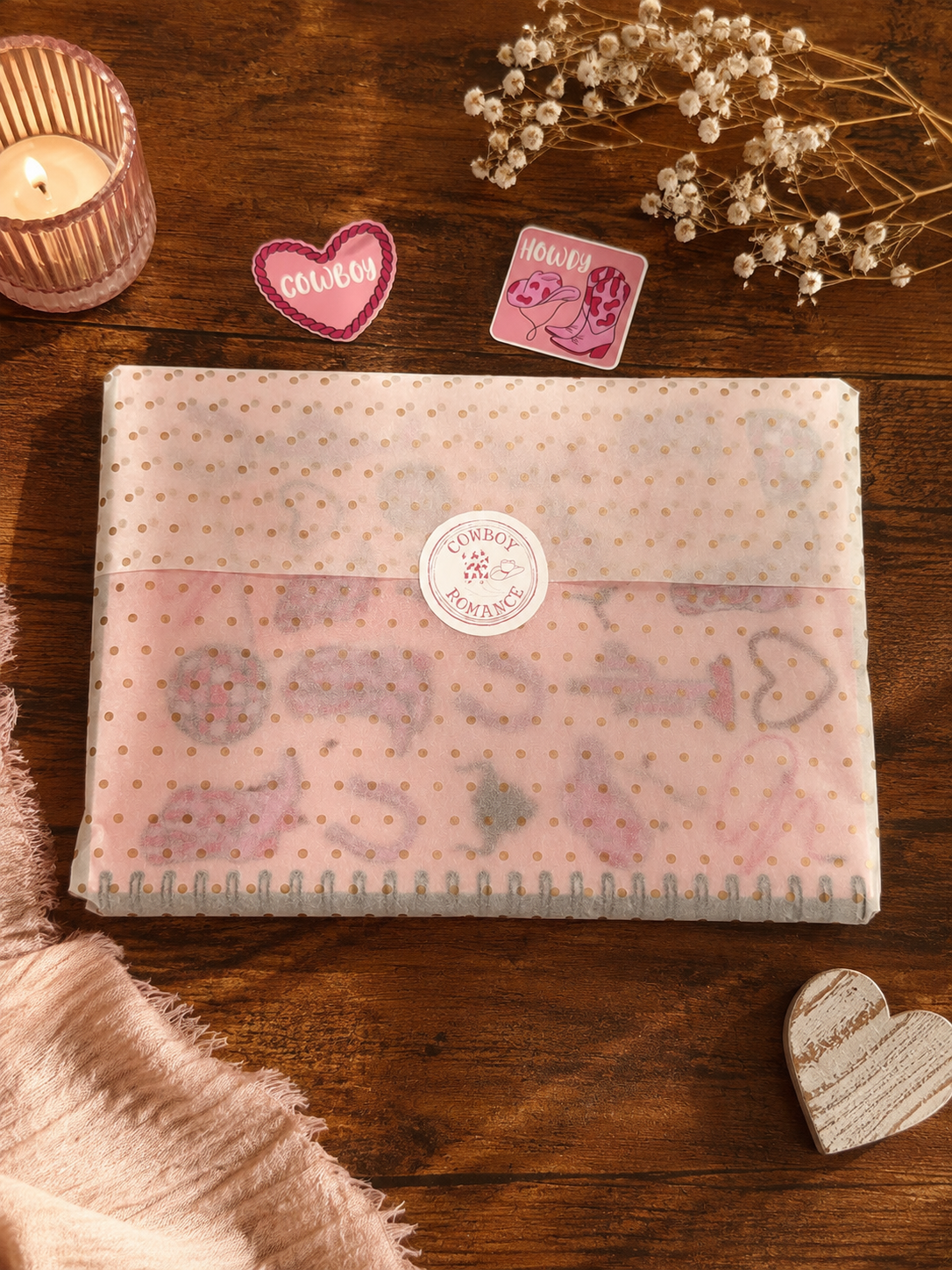 A pastel pink notebook with a dotted pattern and a round cowboy romance label on the cover, styled on a wooden tabletop with a candle, dried flowers, and small heart-shaped decorations.