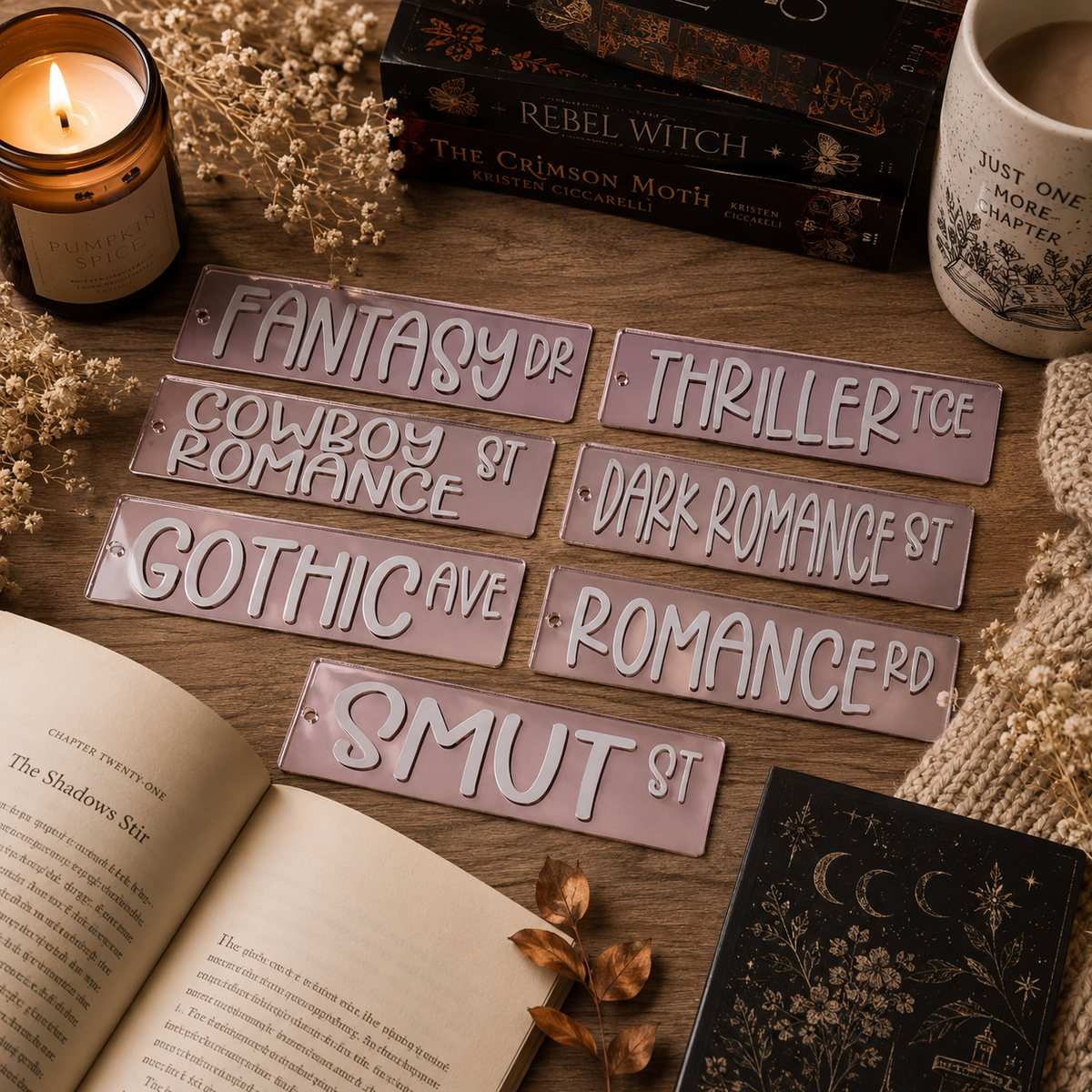 Six rectangular pink bookmark signs with white gothic-style lettering are arranged on a wooden tabletop with candles, books, an open book, dried flowers, and a knitted sweater around them.