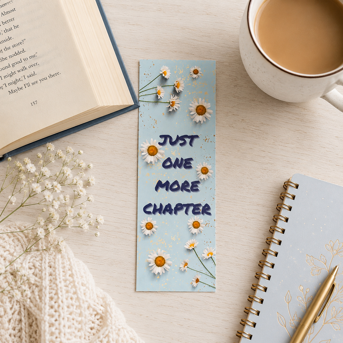 A light blue rectangular book bookmark with daisy illustrations and the text 'JUST ONE MORE CHAPTER' in dark blue, styled on a light wood tabletop with an open book, a cup of coffee, dried flowers, a notebook, and a pen.