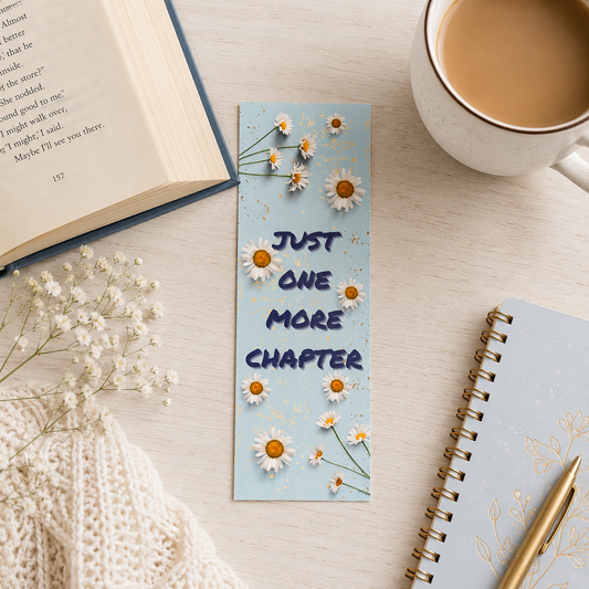 A light blue rectangular book bookmark with daisy illustrations and the text 'JUST ONE MORE CHAPTER' in dark blue, styled on a light wood tabletop with an open book, a cup of coffee, dried flowers, a notebook, and a pen.