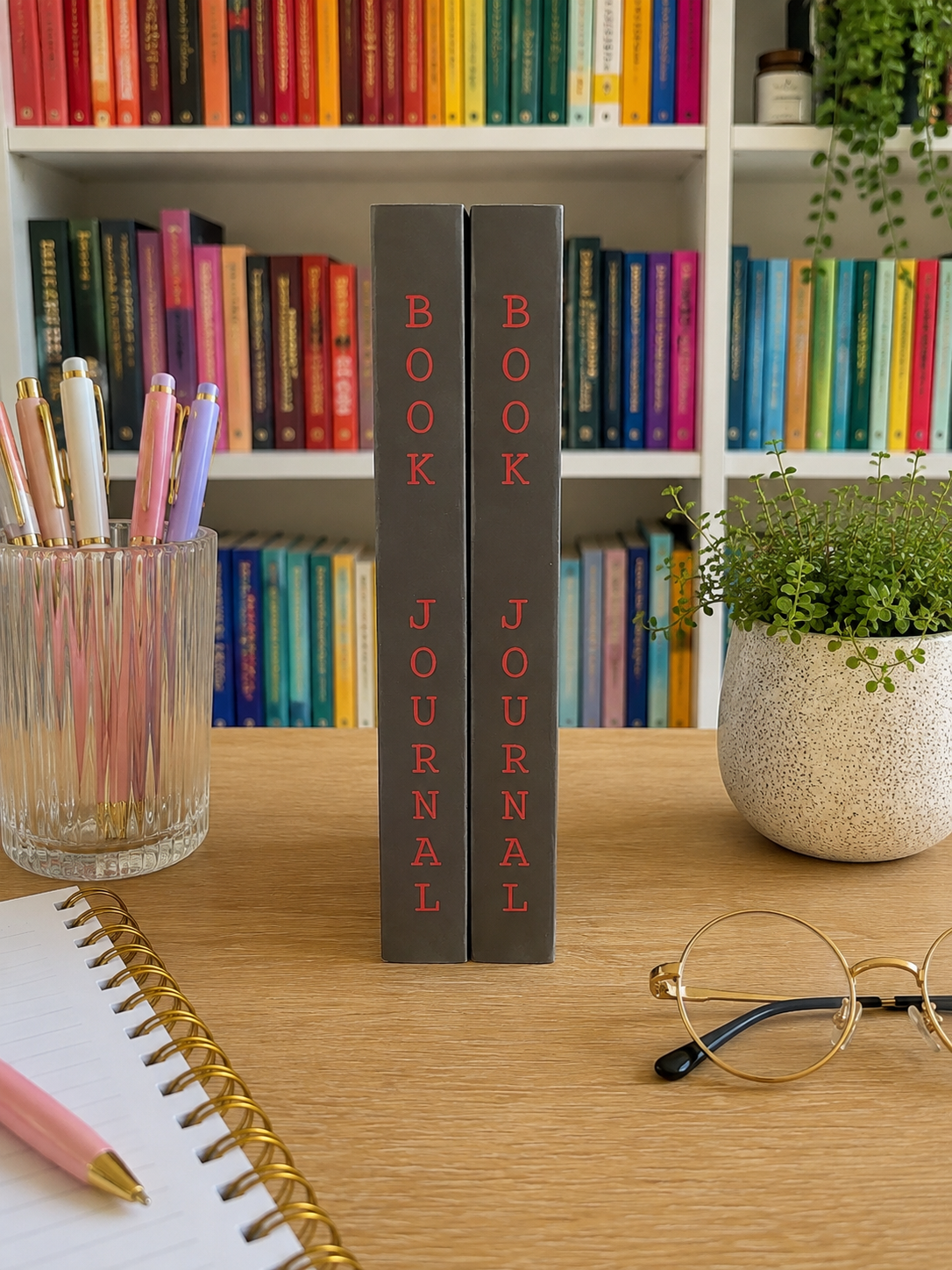 Two slim dark gray dark romance book journals standing upright on a wooden desk, with colorful bookshelves in the background and stationery props around them.
