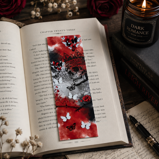 A decorative red and black dark romance themed bookmark with butterflies and roses rests on an open book, styled with a lit candle, books, and a pen in a cozy flat-lay setting.