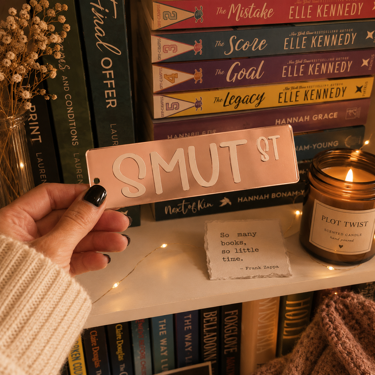 A hand with black nail polish holds a rectangular pink sign that reads 'SMUT ST' in raised cream lettering, positioned in front of a bookshelf with books, a candle, and a small note.