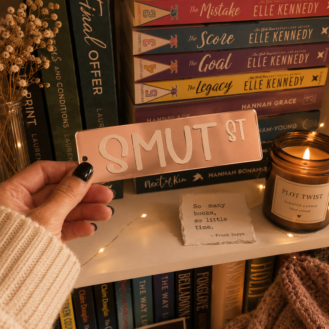 A hand with black nail polish holds a rectangular pink sign that reads 'SMUT ST' in raised cream lettering, positioned in front of a bookshelf with books, a candle, and a small note.