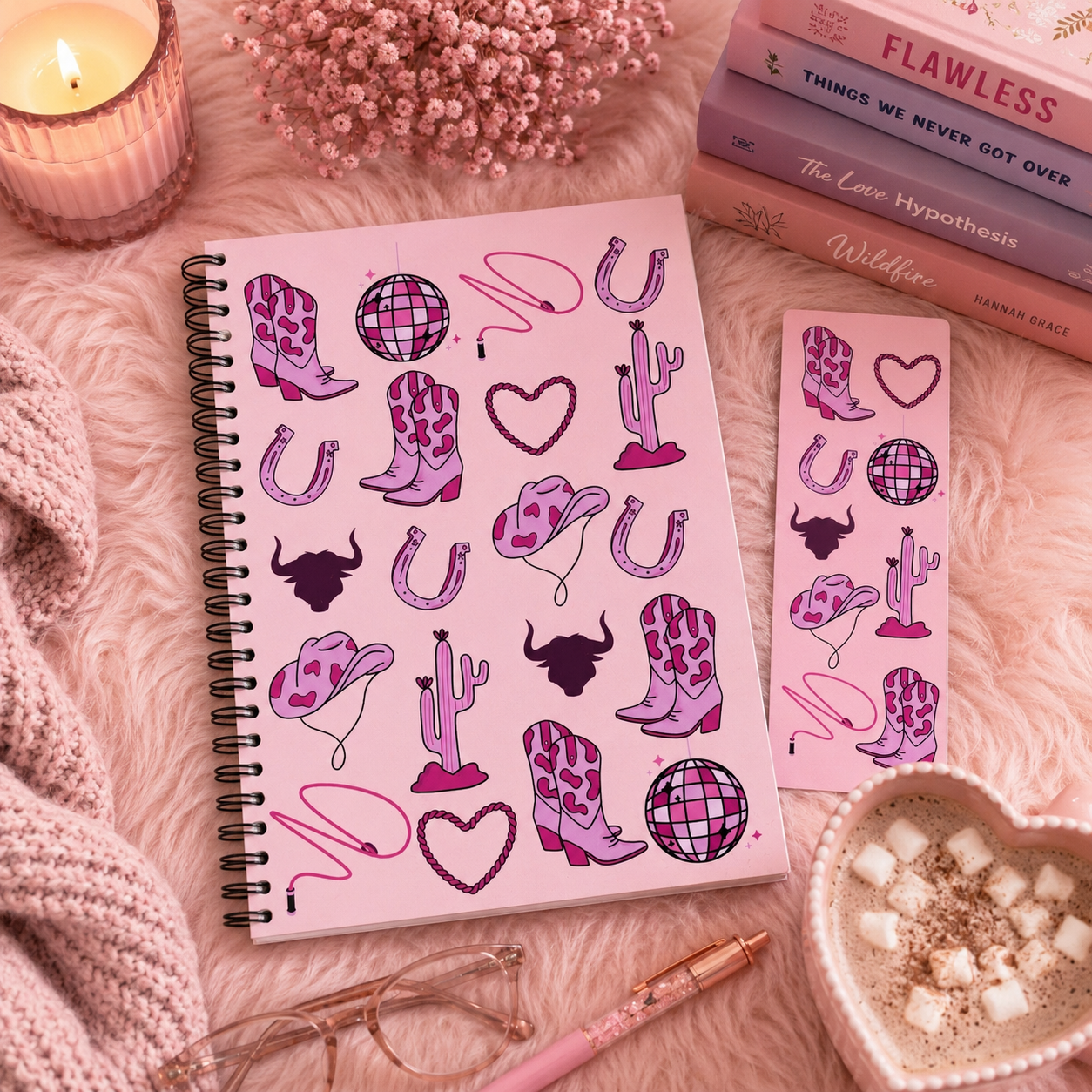 A pink spiral journal and matching sticker sheet featuring cowboy boot, horseshoe, cactus, heart, disco ball, and bowtie illustrations, styled on a fluffy pink blanket with a candle, books, glasses, and a heart-shaped drink bowl.