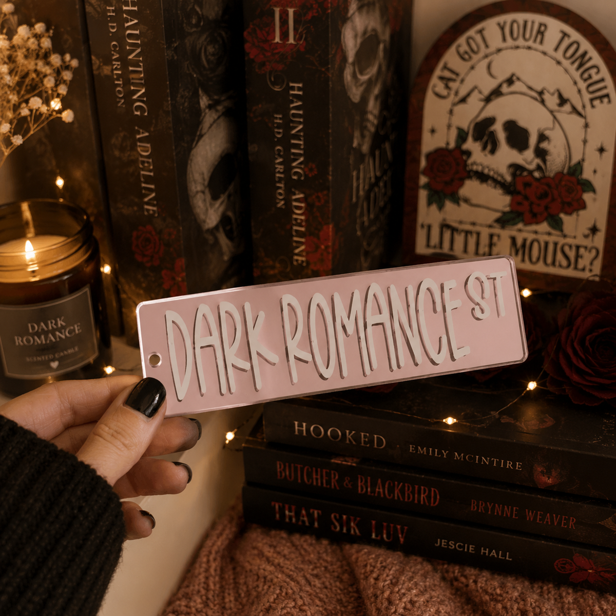 A hand with black nail polish holds a pink rectangular sign that reads 'DARK ROMANCE ST' in white gothic lettering. The background is a cozy, dark-themed bookshelf and room setting with books like 'Haunting Adeline', a lit candle, and a sign reading 'Cat Got Your Tongue? Little Mouse?'.