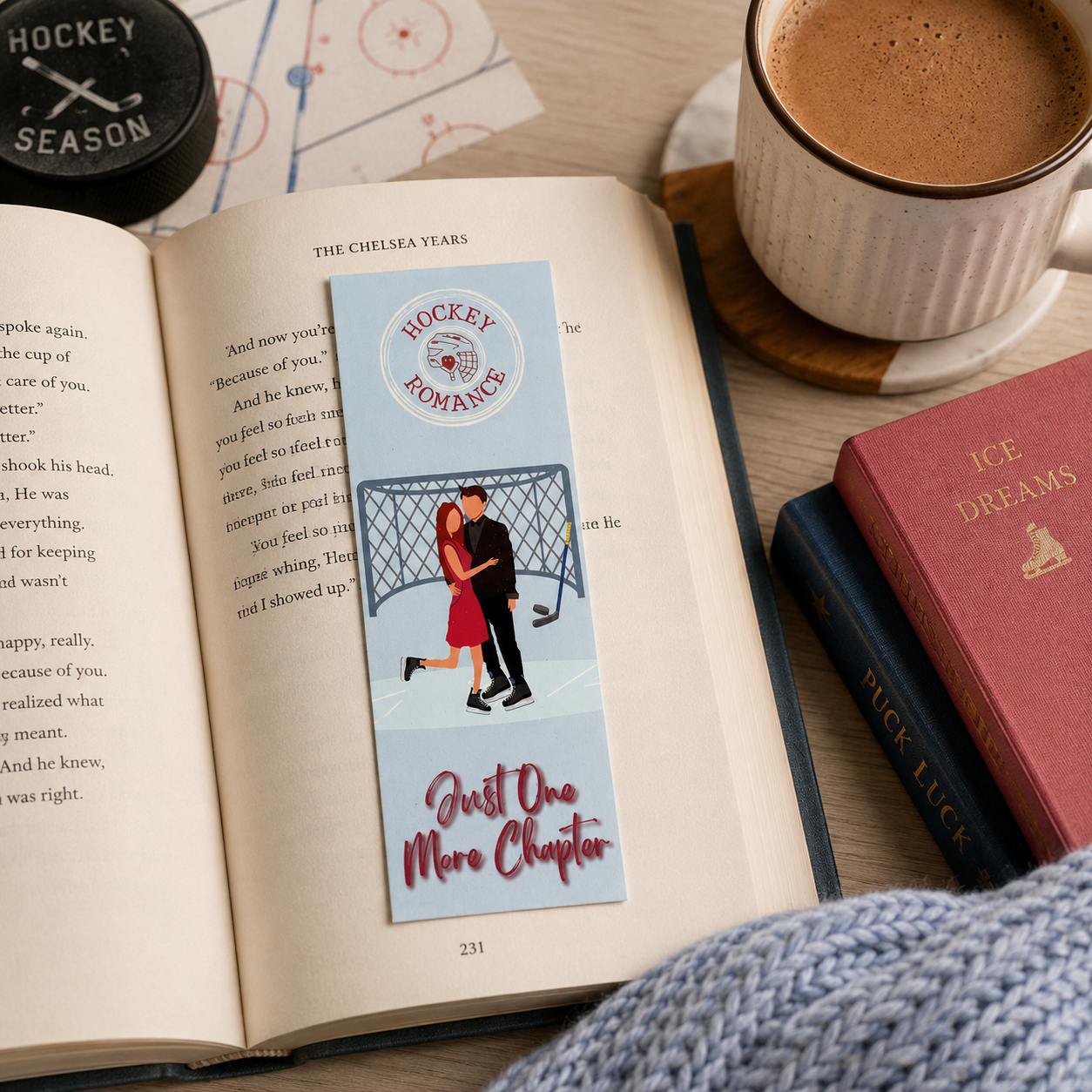 A rectangular book bookmark with a light blue background, featuring a circular 'Hockey Romance' logo and an illustration of a couple in front of a hockey goal, placed on an open book surrounded by a coffee cup, another book, and a knit blanket.
