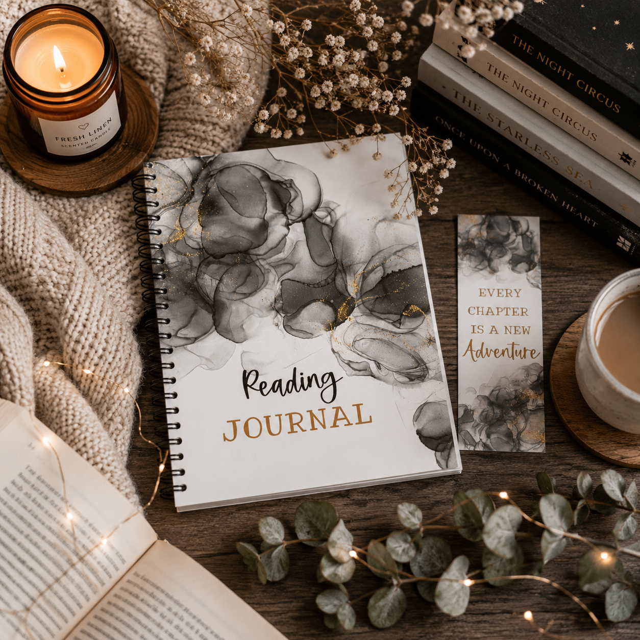 A spiral-bound reading journal with a white cover and grey marble artwork is styled on a wooden surface with a candle, books, a cup of coffee, eucalyptus leaves, fairy lights, and a small bookmark card that reads, 'Every chapter is a new adventure.'.