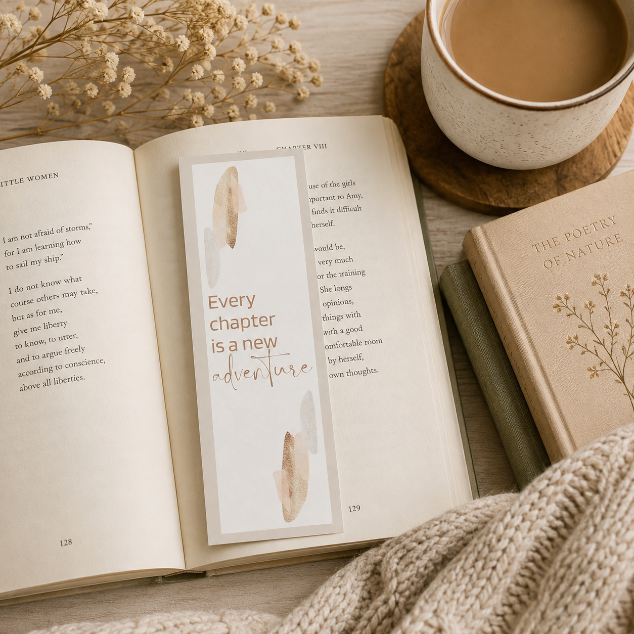 An open book and bookmark spread on a wooden surface with a cream-colored bookmark featuring gold foil text and abstract leaf illustrations. A cup of coffee on a coaster, dried flowers, and a knitted blanket frame the scene.
