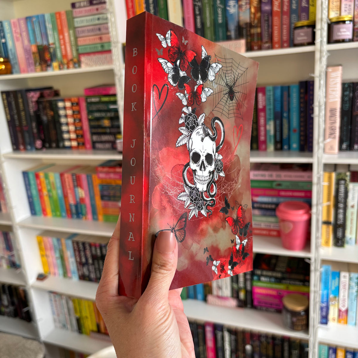 A hand holding a dark romance style reading journal with a red cover featuring a skull and butterflies, in front of a bookshelf filled with various books.
