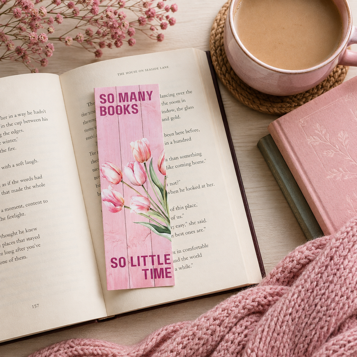 A pink bookmark with a tulip illustration and the text 'SO MANY BOOKS SO LITTLE TIME' placed on an open book, styled in a cozy flat lay with a pink mug, knit blanket, and another book.