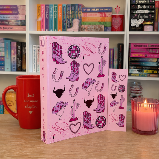 A pink book journal with western-themed illustrations, shown standing on a wooden table with a matching journal page insert, a red mug, and a lit candle, set in front of a bookshelf.