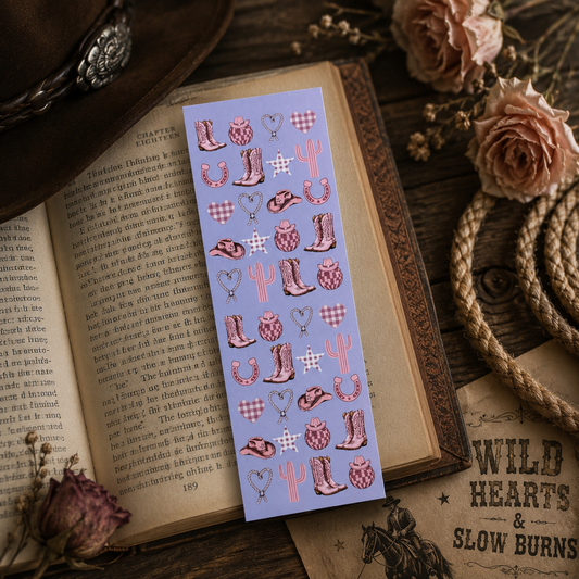 A bookmark with western-themed illustrations, including cowboy boots, cacti, horseshoes, hearts, and plaid patterns, displayed on top of an open vintage book surrounded by dried flowers and rope in a cowboy romance theme