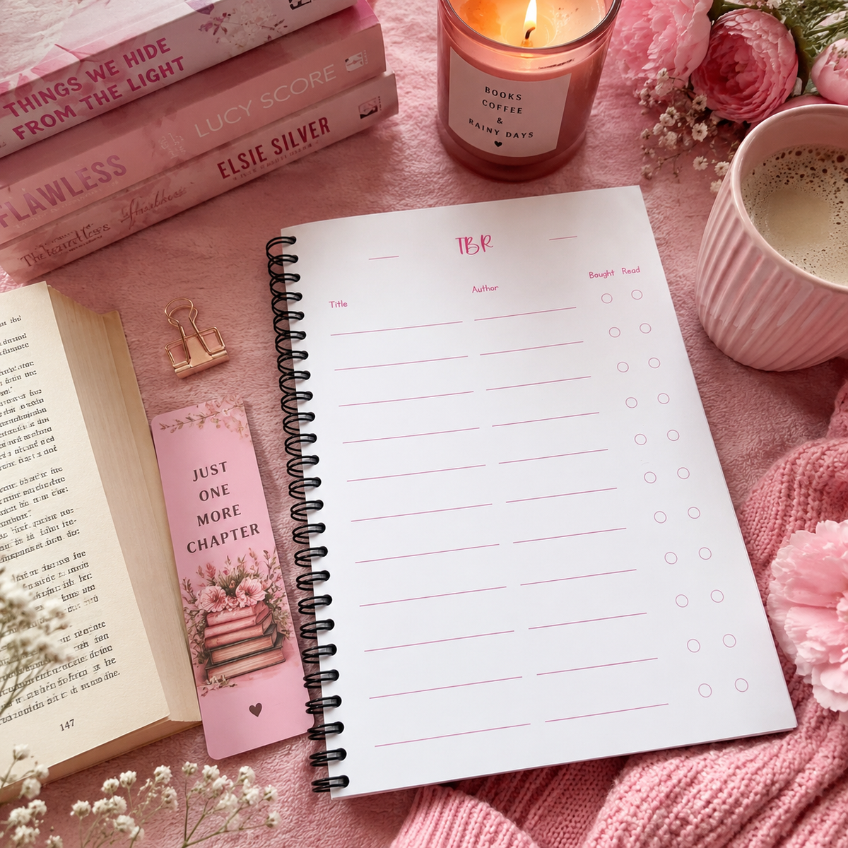 A spiral-bound reading journal page with a pink header reading 'TBR' is arranged on a pink fabric surface with books, a candle, a cup of coffee, flowers, and a pink knit blanket.