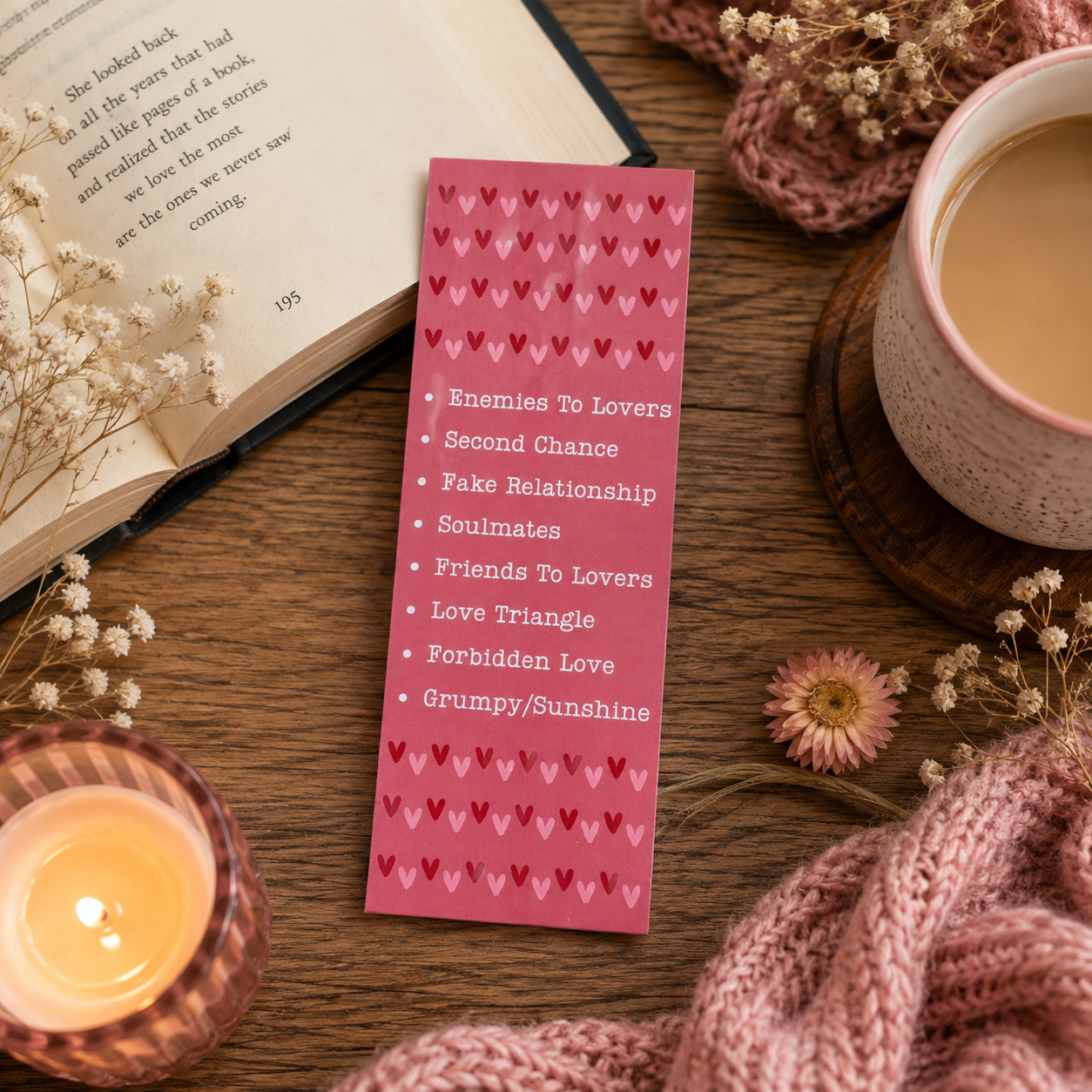 A pink rectangular bookmark with rows of small red hearts and a bulleted list of romantic phrases, styled on a wooden table with an open book, candle, cup of tea, and knitted scarf.