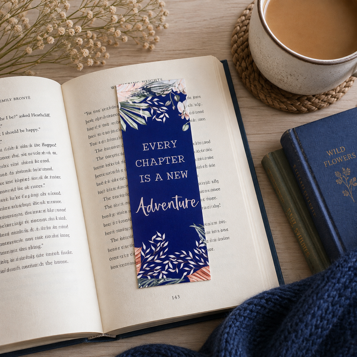 A navy blue book bookmark with white and pink tropical leaf patterns and the text 'EVERY CHAPTER IS A NEW Adventure' lies on an open book on a wooden table, styled with a cup of coffee, a book, and a knit sweater.